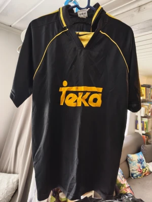 Svart Nike fotbollströja med gula detaljer/xl  - Cool svart fotbollströja från Nike med gula detaljer och Teka-logga på bröstet. Tröjan har korta ärmar, V-ringning med krage och gula sömmar längs axlarna. Perfekt för träning eller match! Slitet märke 