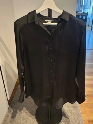 Svart blus från H&M - Stilren svart blus från H&M i ett silkeslent material. Blusen har klassisk krage, knappar framtill och långa ärmar. Perfekt för dig som gillar en enkel men elegant look.