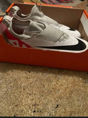 Nike vita fotbollsskor med svart swoosh - Säljer ett par vita Nike fotbollsskor med stor svart swoosh på ovansidan och rosa detaljer på hälen. Skorna har en modern design med täckt snörning och elastisk plös för en tight passform. Perfekta för dig som vill sticka ut på planen.
