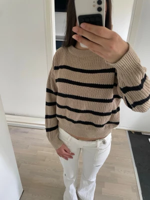 Beige stickad tröja med svarta ränder - Säljer en beige stickad tröja med svarta horisontella ränder. Tröjan har en avslappnad passform, rund halsringning och ribbade muddar vid ärmslut och nederkant. Perfekt för lager på lager och enkel att styla till olika outfits. Storleken är 150 men passar bra till nån som har 170 (xs,s)