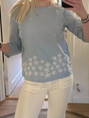 Ljusblå blus med broderade blommor - Supersöt ljusblå blus med vita broderade blommor längs nederkanten. Blusen har trekvartsärm och rak passform. Perfekt att matcha med ljusa jeans för en fräsch look.