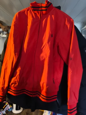 Röd vindjacka från Polo Ralph Lauren - Röd vindjacka med svarta detaljer från Polo Ralph Lauren. Jackan har dragkedja framtill, ribbade muddar och krage med svarta ränder samt broderad logga på bröstet. Perfekt för blåsiga dagar och snygg till streetwear-stilen. Tyvärr är de fläckar på den 