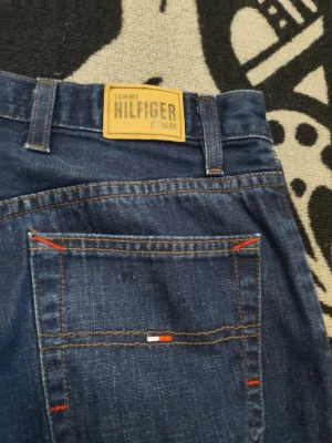 Blå jeans från Tommy Hilfiger Denim - Snygga blå jeans från Tommy Hilfiger Denim med raka ben och klassisk femficksdesign. Jeansen har kontrastsömmar i orange och en läderpatch med logga bak i midjan. Tillverkade i slitstark denim med diskret broderad logga på bakfickan.
