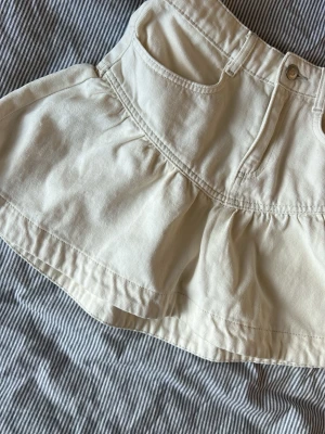 Vit volangkjol - Säljer en beige kortkjol med volang och praktiska fickor framtill. Kjolen har en klassisk jeanslook med knapp och dragkedja i midjan. Perfekt för dig som gillar en avslappnad och trendig stil.