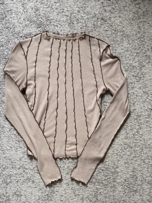 Beige ribbad långärmad topp - Trendig beige ribbad topp med långa ärmar och vågiga kanter. Toppen har bruna vertikala sömmar som ger en cool detalj och croppad passform. Perfekt att styla med jeans eller kjol för en snygg look.