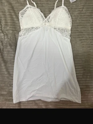Vitt spetsnattlinne med tunna axelband - Elegant vitt nattlinne med tunna justerbara axelband och vacker spetsdetalj över bysten. Materialet är mjukt och lätt, perfekt för en bekväm och stilren look hemma. Liten pärldekoration framtill ger extra charm.