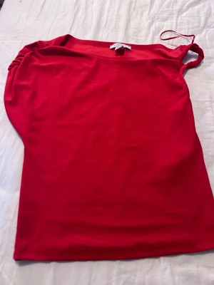 Röd oneshoulder topp  - Trendig röd oneshoulder topp  med snygg rynkdetalj på axeln. Toppens material känns mjukt och stretchigt, perfekt för att skapa en cool och stilren look. Passar dig som gillar att sticka ut med färg och form.