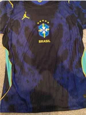 Blå Brasilien fotbollströja Jordan - Snygg blå och svart fotbollströja från Brasilien med CBF-logga och fem stjärnor på bröstet. Tröjan har gula detaljer, blåa och svarta mönster samt gul Jordan-logga. Tillverkad i lätt och ventilerande material, perfekt för träning eller match.