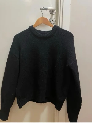 Svart stickad tröja med rund hals - Mysig svart stickad tröja med rund halsringning och långa ärmar. Tröjan har en enkel, oversized passform och ribbade muddar vid ärmslut och nederkant. Perfekt för lager på lager och en avslappnad stil.