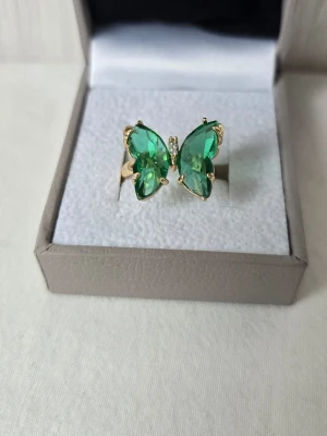 Guldfärgad ring med grön fjäril - Unik guldfärgad ring med en stor fjärilsformad dekoration i gröna kristaller. Fjärilen har detaljer i guld och små klara stenar i mitten som ger extra bling. Perfekt för dig som vill sticka ut med ett färgstarkt och trendigt smycke. Justerbar 