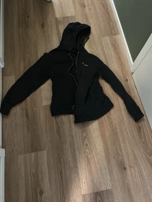 Svart hoodie från Marc O'Polo - Säljer en svart hoodie från Marc O'Polo med dragkedja framtill och vit broderad logga på bröstet. Hoodien har huva med dragsko och ribbade muddar vid ärmslut och nederkant. Perfekt för en avslappnad och stilren look.