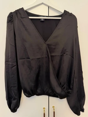 Svart omlottblus från Sincerely Jules - Stilren svart omlottblus från Sincerely Jules med djup V-ringning och resår i midjan. Blusen har långa puffärmar och är tillverkad i ett glansigt, mjukt material som ger en lyxig känsla. Perfekt att styla med både jeans och kjol.
