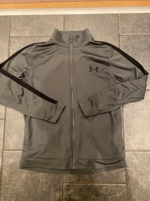 Grå ziptröja från Under Armour - Snygg grå ziptröja från Under Armour med svarta detaljer längs ärmarna och diskret logga på bröstet. Tröjan har hög krage, hel dragkedja och är tillverkad i ett mjukt, stretchigt funktionsmaterial som passar perfekt till träning.