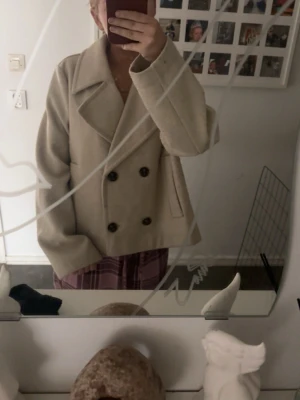 Beige dubbelknäppt kappa från Vero Moda - Säljer en beige kort kappa från Vero Moda med breda slag och dubbelknäppning. Kappan har två sidofickor och är tillverkad i ett ull-liknande material som ger en stilren och klassisk look. Perfekt för att höja din outfit under kyliga dagar.
