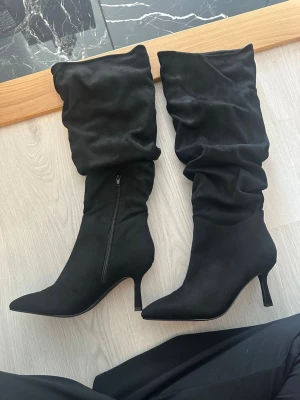 Svarta stövlar med klack - Säljer ett par svarta stövlar med slouchy skaft och spetsig tå. De har smal klack och dragkedja på insidan. Materialet är mockaimitation som ger en mjuk och lyxig känsla. Perfekta för att lyfta vilken outfit som helst.