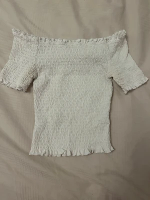 Vit offshoulder topp med volang från H&M - Vit offshoulder topp med korta ärmar och volangdetaljer vid kanterna från H&M i storlek XS. Smockad och stretchig passform som sitter snyggt på kroppen. 