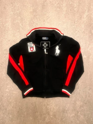 Polo Ralph Lauren full zip - Svart tröja med dragkedja från Polo Ralph Lauren. Tröjan har röda och vita detaljer, broderade märken på bröstet och Schweiz-flagga samt siffran 10 på ryggen. Sportig design med ståkrage och röda ränder längs ärmarna.
