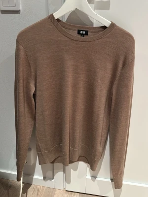 Beige stickad tröja från Uniqlo - En stilren beige stickad tröja från Uniqlo med rund halsringning och långa ärmar. Tröjan har en tunn och mjuk kvalitet, perfekt för lager på lager. Ribbstickade muddar vid ärmslut och nederkant ger en snygg finish. 100% ull