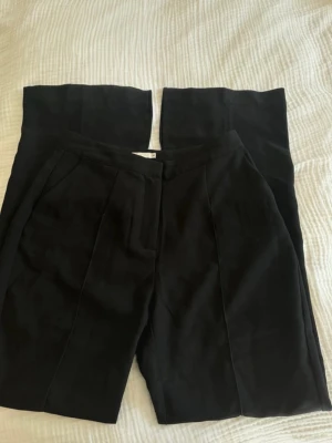 Svarta vida kostymbyxor med slits - Säljer ett par svarta kostymbyxor med hög midja och vida ben. Byxorna har pressveck framtill och sidofickor för en clean look. Perfekta till en snygg outfit när du vill känna dig extra uppklädd.