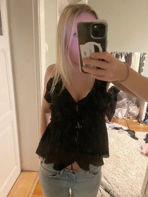Svart volangblus med snörning - Säljer en svart blus med volanger och snörning framtill. Blusen är ärmlös och har en v-ringad design som ger en snygg och trendig look. Perfekt att matcha med jeans eller kjol för en cool stil.