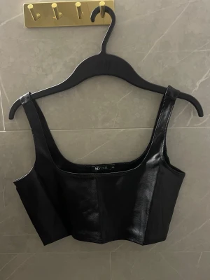 Svart korsettopp i skinnimitation - Snygg svart korsettopp från NLY ONE i skinnimitation. Toppen är croppad med breda axelband och har en stilren, figurnära passform. 