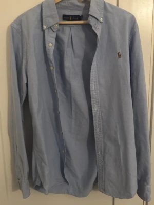 Ljusblå skjorta från Ralph Lauren - Klassisk ljusblå skjorta från Ralph Lauren med broderad logga på bröstet. Skjortan har lång ärm, knappar framtill och button-down krage. Tillverkad i mjuk bomull som känns skön mot huden. Perfekt för en clean och stilren look.