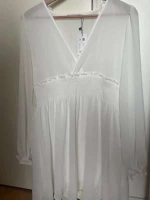 Vit omlottklänning med smock - Söt vit klänning från Lager 157, med omlott-ringning och smockad midja. Klänningen har långa, lätt transparenta ärmar med resår vid ärmslut och ett luftigt fall. Perfekt för en fräsch och stilren look. Kan sänka priset!!