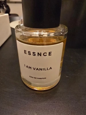 ESSNCE I Am Vanilla Eau de Parfum - En stilren parfymflaska från ESSNCE med doften I Am Vanilla. Flaskan är genomskinlig med en vit etikett och svart text, och har ett svart lock. Innehållet är gyllene och flaskan är tillverkad i glas.