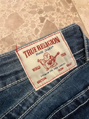 True Religion Joey Low Rise Flare Jeans - Blå jeans från True Religion i modellen Joey Low Rise Flare. Jeansen har låg midja, utsvängda ben och fickor med lock. Tillverkade i klassisk denim med synliga kontrastsömmar och cool vintagekänsla. Perfekt för dig som gillar en trendig och avslappnad look. Oanvända. Lappen kvar. Nypris 1299kr