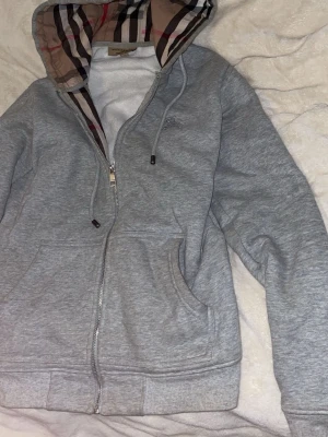 Grå zip hoodie från Burberry - Säljer en grå hoodie från Burberry med dragkedja och klassiskt rutigt foder i huvan. Hoodien har fickor framtill och ribbade muddar. Perfekt för dig som gillar stilrena plagg med exklusiv känsla och snygga detaljer.