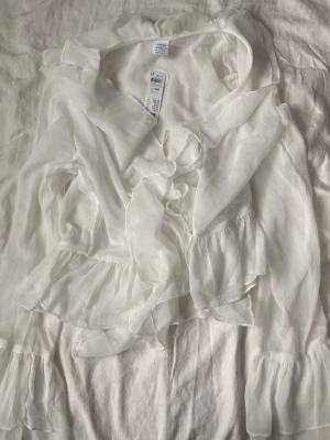 Vit blus - Superfin vit blus från Lindex med volangdetaljer och knytning fram. Blusen har långa ärmar med volang vid ärmslut och är tillverkad i ett lätt, transparent tyg som ger en luftig känsla. Perfekt för en trendig och romantisk look.