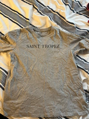 Grå t-shirt med tryck Saint Tropez - Snygg grå t-shirt med svart texttryck 'SAINT TROPEZ' på bröstet. Klassisk rund halsringning och korta ärmar. Mycket bra skick och använd otroligt varsamt. 90% bomull & 10% viskos.