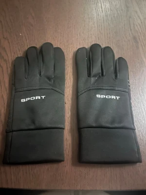 Svarta sporthandskar med grepp - Svarta sporthandskar med tryckt text 'SPORT' på ovansidan. Handskarna har greppmönster på insidan av handflatan och fingertopparna, vilket gör dem perfekta för utomhusaktiviteter. Tillverkade i syntetmaterial och har en stilren, enkel design.