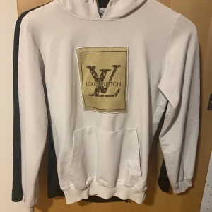Vit Louis Vuitton hoodie med logga - Vit hoodie från Louis Vuitton med stor LV-logga i guld och brunt framtill. Klassisk huva, magficka och ribbade muddar. Skön och stilren, perfekt för dig som vill sticka ut med exklusiv streetstyle.