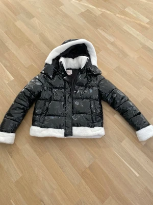 Svart glansig pufferjacka med fuskpäls - Säljer en svart glansig pufferjacka från gina tricot med vit fuskpäls i huva, krage och ärmslut. Jackan har dragkedja och tryckknappar framtill samt en mysig, vadderad känsla. 