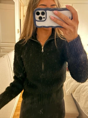 Svart stickad tröja med halvzip - En svart stickad tröja med ribbad struktur och halvzip framtill. Tröjan har hög krage och långa ärmar, perfekt för att hålla värmen. Passar dig som gillar en enkel och stilren look.