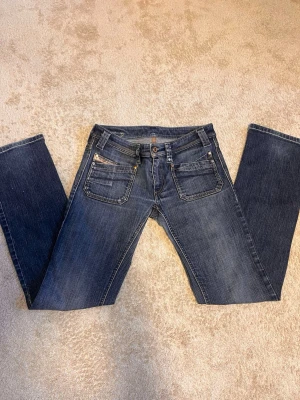 Mörkblå lågmidjade  bootcut jeans med detaljer - Säljer ett par mörkblå bootcut jeans med snygga slitningar och kontrastsömmar. Jeansen har fem fickor, dekorativa nitar och en liten logga på bakfickan. Klassisk knappgylf och cool tvätt som ger en avslappnad vibe.
