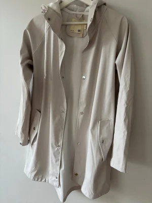 Ljusbeige regnjacka från 157 - Stilren ljusbeige regnjacka från 157 med huva och dragsko. Jackan har tryckknappar framtill, två stora fickor med lock och långa ärmar. Materialet är vattenavvisande och jackan har en rak passform som passar perfekt för regniga dagar.