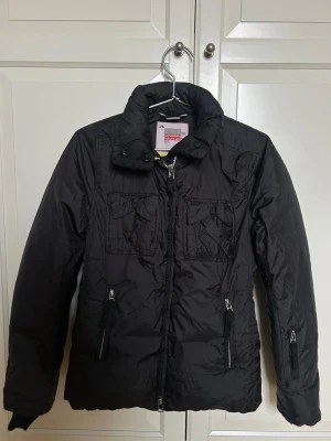 Svart pufferjacka från J.Lindeberg - Snygg svart pufferjacka från J.Lindeberg Future Sports med hög krage och dragkedja framtill. Jackan har två bröstfickor och två sidofickor med dragkedja. Tillverkad i ett glansigt syntetmaterial som håller värmen under kalla dagar. Den är använd 1-2 gånger.