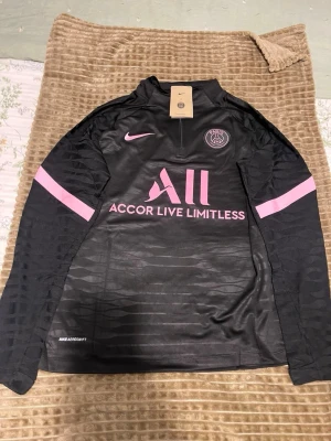 Svart PSG träningsset från Nike - Säljer ett svart träningsset från Nike med PSG-logga, bestående av långärmad tröja och matchande byxor. Setet har rosa detaljer och tryck, inklusive Nike swoosh och Accor Live Limitless. Materialet är lätt och andas, perfekt för sport och träning.