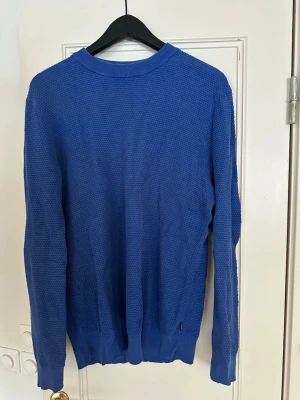Blå stickad tröja med struktur - En stilren blå stickad tröja med diskret strukturmönster. Tröjan har rund halsringning, långa ärmar och ribbade muddar vid ärmslut och nederkant. Perfekt för lager på lager och enkel att matcha med jeans eller chinos. 
