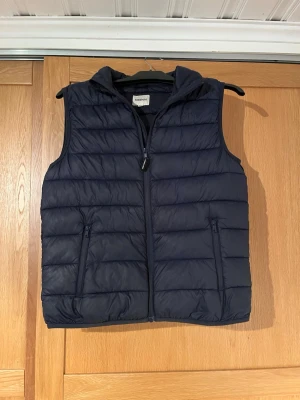 Mörkblå dunväst med huva - Snygg mörkblå dunväst från Kappahl med dragkedja framtill. Västen har två fickor med dragkedja och quiltad design som ger en sportig look. Perfekt att slänga över en hoodie eller tröja för extra värme under kyliga dagar. Tyvärr så har jag behövt laga den på ryggen men det syns inte alls något när man har på sig den och inget man tänker på.