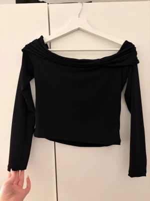Svart offshoulder topp med långa ärmar - Trendig svart offshoulder topp med armbågs långa ärmar och snyggt vikt kant upptill. Toppen har en croppad passform och är tillverkad i ett mjukt, stretchigt material som sitter bekvämt. Perfekt att styla med höga jeans eller kjol.