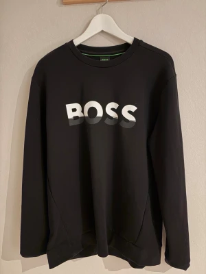 Svart hoodie från BOSS med logga - Svart tjocktröja från BOSS med stor vit logga på bröstet med skuggeffekt. Klassisk rund halsringning och långa ärmar. 