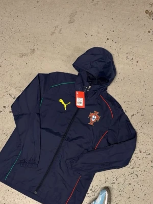 Mörkblå Portugal vindjacka Puma - Snygg mörkblå vindjacka från Puma med Portugals landslagsemblem på bröstet. Jackan har huva, hel dragkedja och detaljer i gult, grönt och rött. Perfekt för dig som vill ha sportig stil och visa ditt stöd för Portugal.