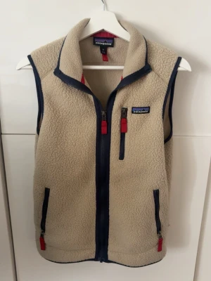 patagonia väst - Säljer en beige fleeceväst från Patagonia med marinblå detaljer och dragkedjor med röda dragare. Västen har hög krage, tre fickor med dragkedja och är tillverkad i mjukt teddyfleece-material. Perfekt för lager-på-lager och outdoor-vibe.