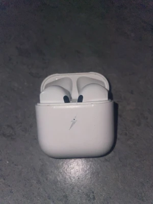 Trådlösa vita earbuds med laddningsetui - Säljer ett par trådlösa vita earbuds med tillhörande laddningsetui. Hörlurarna har en stilren design och etuiet har en blixtsymbol på framsidan. Perfekta för musik, poddar och samtal på språng.