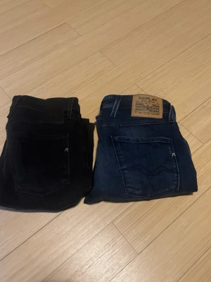 Replay jeans i svart och blå denim - Två par jeans från Replay, ett par i klassisk blå denim med brun läderpatch och ett par i helsvart. Båda har raka bakfickor med diskret R-logga. Perfekta för dig som gillar stilrena och tidlösa jeans i mjukt bomullsmaterial.
