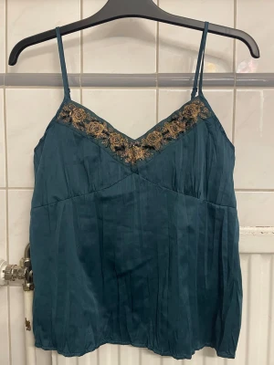Mörkgrön linnetopp med spetsdetalj - Snygg mörkgrön linnetopp med tunna axelband och v-ringning. Toppen har dekorativ spets i svart och guld längs ringningen och är tillverkad i ett glansigt, silkigt material som ger en lyxig känsla. Perfekt att styla med jeans eller kjol.