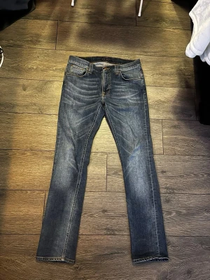 Blå jeans från Nudie Jeans - Säljer ett par klassiska blå jeans från Nudie Jeans med raka ben och snygga slitningar. Jeansen har fem fickor, orange sömmar och den ikoniska Nudie-bågen på bakfickan. Tillverkade i slitstark denim, perfekta för en avslappnad stil.